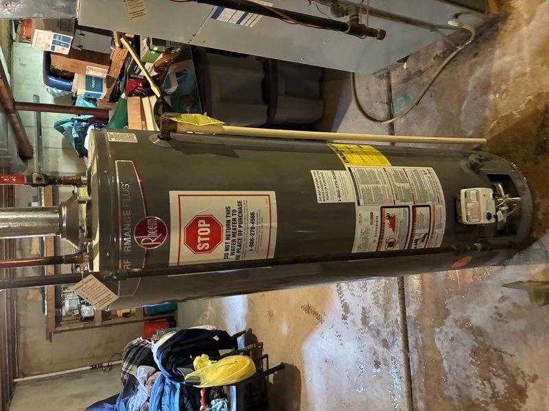 Completed inspection and documentation of tanked water heater installation in residential basement utility room. Unit is a standard vertical gray cylindrical water heater with manufacturer safety labels and technical specifications properly displayed. Verified proper positioning of equipment with adequate clearance for maintenance access. Documented surrounding mechanical room conditions including concrete flooring, exposed ductwork, and HVAC system integration. All safety warning labels including red STOP notifications are clearly visible and compliant with code requirements.