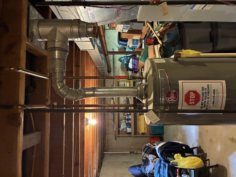 Completed inspection and documentation of tanked water heater installation in residential basement utility room. Unit is a standard vertical gray cylindrical water heater with manufacturer safety labels and technical specifications properly displayed. Verified proper positioning of equipment with adequate clearance for maintenance access. Documented surrounding mechanical room conditions including concrete flooring, exposed ductwork, and HVAC system integration. All safety warning labels including red STOP notifications are clearly visible and compliant with code requirements.