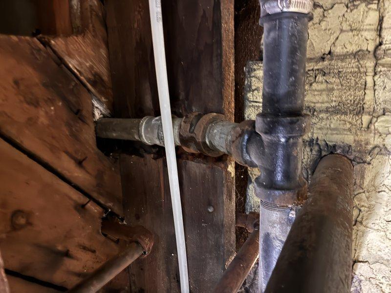 # Clogged Drain Service - Plumbing System Inspection and Assessment

Responded to a clogged drain call and conducted a thorough inspection of the plumbing system in the attic/crawl space area. Upon investigation, examined the PVC drainage piping configuration, including T-junction connections and vertical drain lines secured with metal hose clamps. Inspected the condition of both white PVC pipes and older galvanized metal piping showing signs of age and oxidation. Evaluated the flexible rubber drainage hose connections secured with metal clamps to the main PVC system. Assessed the overall drainage assembly routing through wooden structural beams and verified proper mounting with pipe straps. Documented the plumbing infrastructure condition, including the presence of spray foam insulation sealing gaps around pipe penetrations. System components were photographed for service records, showing the drainage configuration and connection points throughout the confined utility space.