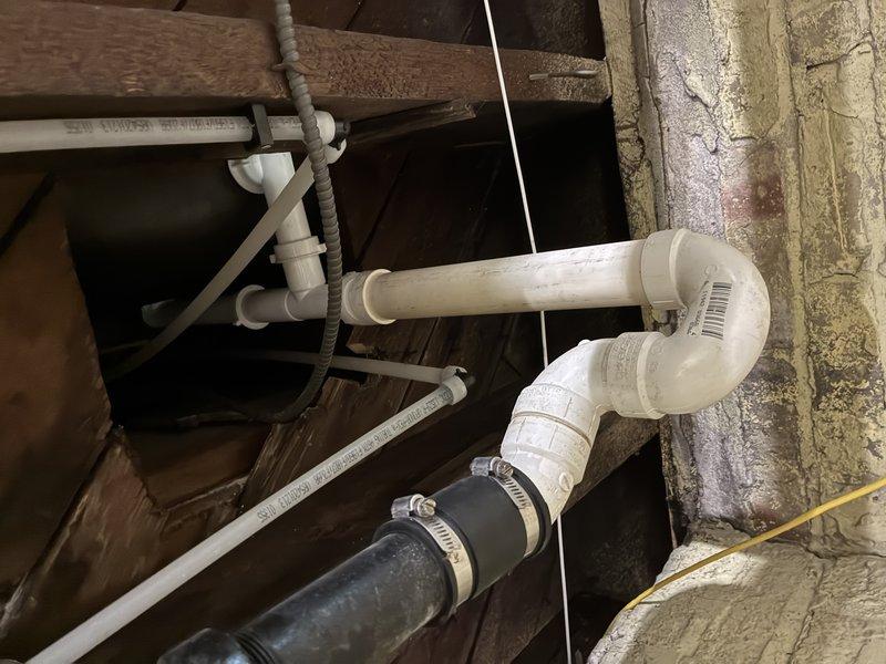 # Clogged Drain Service - Plumbing System Inspection and Assessment

Responded to a clogged drain call and conducted a thorough inspection of the plumbing system in the attic/crawl space area. Upon investigation, examined the PVC drainage piping configuration, including T-junction connections and vertical drain lines secured with metal hose clamps. Inspected the condition of both white PVC pipes and older galvanized metal piping showing signs of age and oxidation. Evaluated the flexible rubber drainage hose connections secured with metal clamps to the main PVC system. Assessed the overall drainage assembly routing through wooden structural beams and verified proper mounting with pipe straps. Documented the plumbing infrastructure condition, including the presence of spray foam insulation sealing gaps around pipe penetrations. System components were photographed for service records, showing the drainage configuration and connection points throughout the confined utility space.