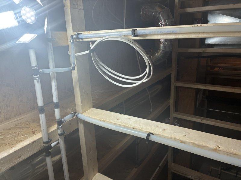 Completed installation of white PVC drainage piping system within exposed wooden framework of unfinished interior space. Installed vertical pipe runs secured with metal brackets and straps to structural framing, including proper fitting connections and flexible hose routing through utility area to ensure code-compliant drainage configuration.