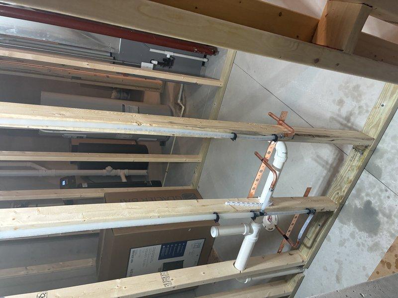 Completed installation of white PVC drainage piping system within exposed wooden framework of unfinished interior space. Installed vertical pipe runs secured with metal brackets and straps to structural framing, including proper fitting connections and flexible hose routing through utility area to ensure code-compliant drainage configuration.