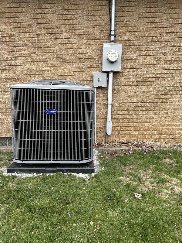 # HVAC Install - Complete System

## Summary
Completed full HVAC system installation including outdoor Carrier condenser unit with electrical service components and indoor mechanical equipment with associated connections and controls.

## Work Performed

**Outdoor Equipment Installation:**
Installed Carrier brand air conditioning condenser unit on prepared black concrete pad against exterior brick wall. Positioned unit to ensure proper clearance and accessibility for future maintenance. Mounted electrical disconnect box and meter socket on brick wall with white electrical conduit running vertically to service the outdoor unit. Secured all electrical connections per manufacturer specifications and local electrical codes. Verified proper grounding and bonding of all electrical components.

**Indoor Equipment Installation:**
Installed cylindrical mechanical equipment in basement utility area with proper clearance from walls and surrounding structures. Connected multiple refrigerant lines 
