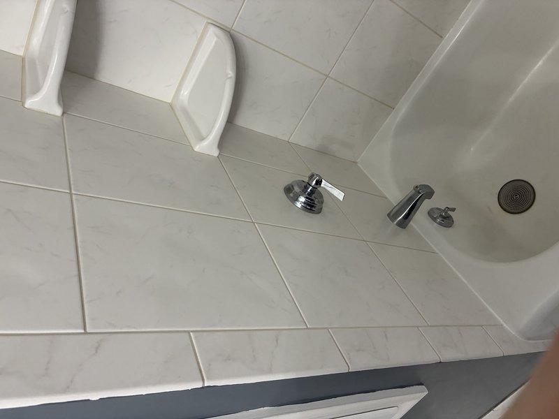 Completed installation of modern shower system featuring chrome-finished fixtures including wall-mounted showerhead, handheld spray attachment with mounting pole, and control valve assembly. Installed light gray rectangular tiles with marble veining in horizontal brick pattern throughout shower walls and basin area, along with white accessibility grab bar for safety compliance. All fixtures properly mounted and sealed to ensure water-tight operation.