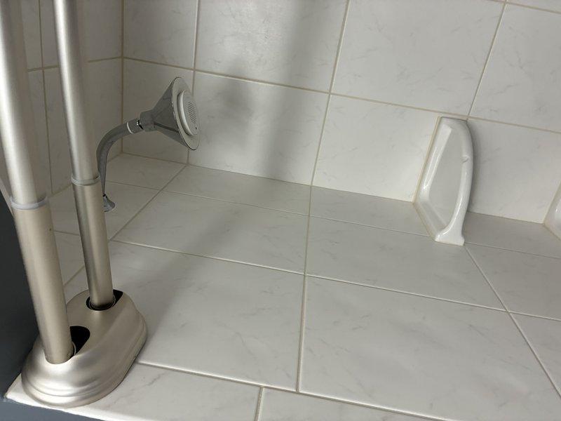 Completed installation of modern shower system featuring chrome-finished fixtures including wall-mounted showerhead, handheld spray attachment with mounting pole, and control valve assembly. Installed light gray rectangular tiles with marble veining in horizontal brick pattern throughout shower walls and basin area, along with white accessibility grab bar for safety compliance. All fixtures properly mounted and sealed to ensure water-tight operation.
