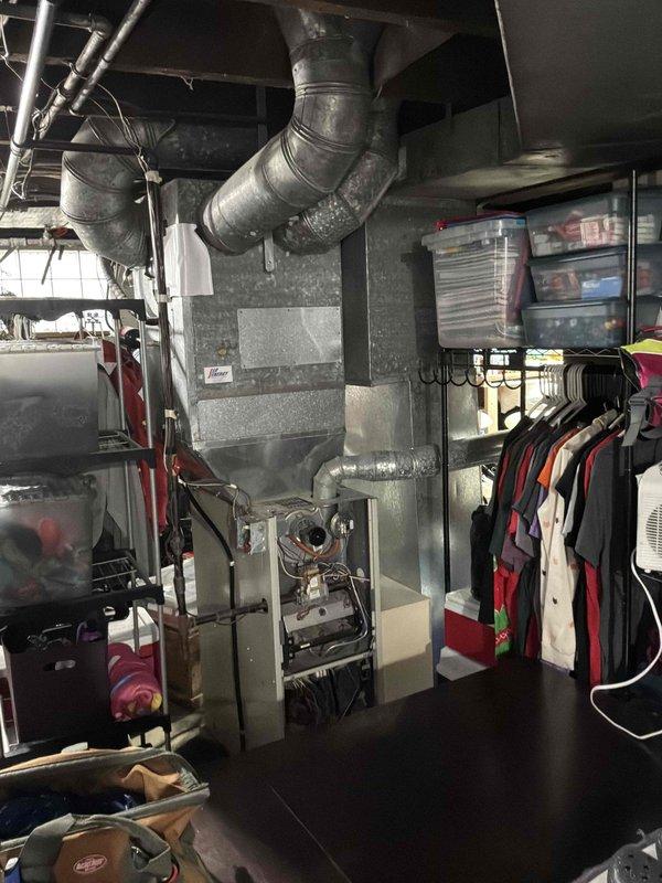 Completed comprehensive HVAC system evaluation in residential basement utility space for furnace replacement estimate. Assessed existing gray metal furnace unit with exposed access panel, documented current ductwork configuration including large metallic supply ducts extending from unit across ceiling, and evaluated spatial constraints for new equipment installation given storage items and recreational equipment in surrounding area. Photographed existing system layout and mechanical components to provide accurate replacement specifications and ensure proper sizing for new high-efficiency heating system.