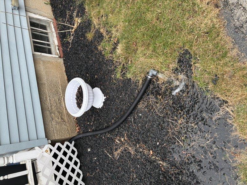 Performed inspection and assessment of existing sump pump system. Upon examination, found sump pump displaying significant corrosion, rust, and mineral deposit buildup on metal components, indicating extended service life and exposure to moisture. Pump was operational but showing clear signs of deterioration that could lead to future failure. Documented condition of circular ribbed plastic sump pit liner and verified proper water level within basin. Inspected white PVC discharge piping connection at pump assembly and traced exterior discharge line. Exterior inspection revealed black corrugated flexible drainage hose routing water away from foundation to appropriate drainage area at lawn perimeter. Verified discharge system was properly directing water away from structure to prevent foundation issues. Photographed all system components for maintenance records. Recommended monitoring pump performance and considering replacement due to advanced corrosion to prevent potential emergency fai