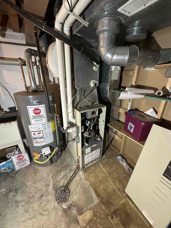 # HVAC System Evaluation and Replacement Estimate

Conducted comprehensive assessment of existing residential HVAC system located in basement utility area. Evaluated current furnace/air handler unit with connected ductwork, including gray metal trunk lines showing dust accumulation and signs of age. Inspected Aprilaire humidifier unit mounted to furnace system and assessed its integration with existing equipment.

Examined water heater installation proximity to HVAC equipment to ensure adequate clearance requirements for replacement unit specifications. Documented existing PVC condensate drainage system, white PVC venting configuration, and black flexible ductwork connections. Assessed copper and brass piping connections at water heater and their potential impact on equipment access during replacement procedures.

Photographed existing equipment layout including Y-shaped ductwork connections extending to ceiling distribution system. Noted space constraints due to stored materials and p