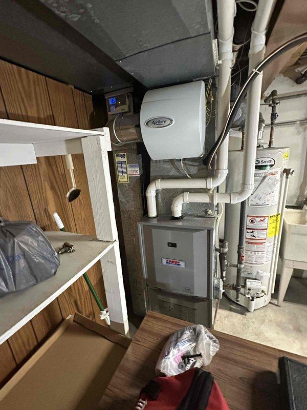 # HVAC System Evaluation and Replacement Estimate

Conducted comprehensive assessment of existing residential HVAC system located in basement utility area. Evaluated current furnace/air handler unit with connected ductwork, including gray metal trunk lines showing dust accumulation and signs of age. Inspected Aprilaire humidifier unit mounted to furnace system and assessed its integration with existing equipment.

Examined water heater installation proximity to HVAC equipment to ensure adequate clearance requirements for replacement unit specifications. Documented existing PVC condensate drainage system, white PVC venting configuration, and black flexible ductwork connections. Assessed copper and brass piping connections at water heater and their potential impact on equipment access during replacement procedures.

Photographed existing equipment layout including Y-shaped ductwork connections extending to ceiling distribution system. Noted space constraints due to stored materials and p