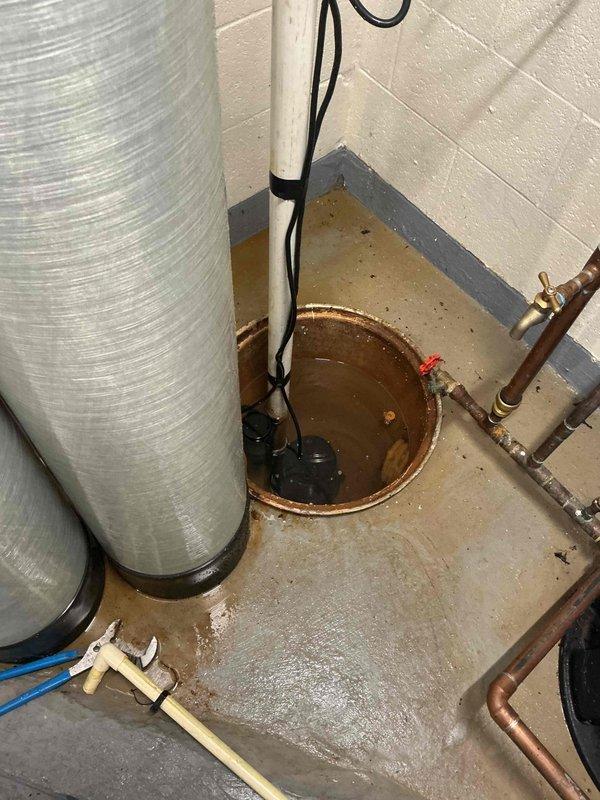 Performed inspection and maintenance of residential sump pump system located in basement utility area. Documented existing sump pump installation within concrete basin, noting rust staining on basin walls and water presence indicating active use. Verified pump operation and assessed discharge piping configuration, including white PVC discharge line. Photographed complete system setup including connection points to ensure proper documentation of equipment condition and plumbing connections for service records.