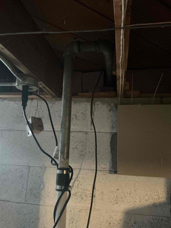 Conducted plumbing estimate for pump system evaluation in basement utility area. Inspected aged vertical discharge pipe showing significant oxidation and weathered patina, assessed electrical connections and wiring runs associated with existing pump installation. Examined exterior foundation perimeter where debris accumulation near building corner may impact drainage systems and potential pump discharge lines, noted need for clearance to ensure proper water evacuation from pump operations.