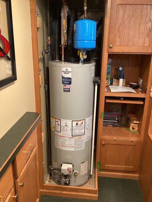 Completed installation of Bradford White residential water heater with blue expansion tank in utility cabinet space. Unit installed on wooden platform base with copper piping connections, brass fittings, and proper safety compliance labeling. Expansion tank mounted above water heater with vertical copper pipe runs and valve connections for pressure regulation in accordance with standard residential plumbing code requirements.