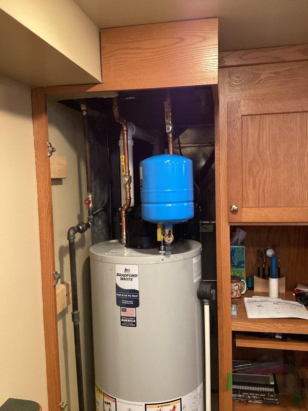 Completed installation of Bradford White residential water heater with blue expansion tank in utility cabinet space. Unit installed on wooden platform base with copper piping connections, brass fittings, and proper safety compliance labeling. Expansion tank mounted above water heater with vertical copper pipe runs and valve connections for pressure regulation in accordance with standard residential plumbing code requirements.