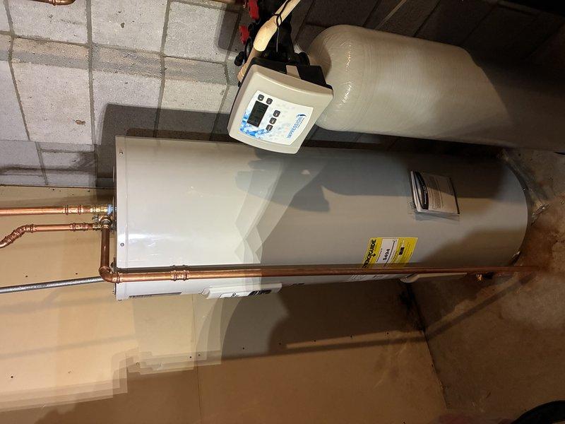 # Plumbing Install - Tanked Water Heater

Completed installation of residential tanked water heater in basement utility area. Installed new cylindrical water heater tank on concrete floor with proper positioning and clearances. Connected hot and cold water supply lines using copper piping with professional compression fittings and joints, forming proper loop configuration at top of unit. Installed brass shut-off valves on both supply lines for isolation and maintenance access. Connected gray galvanized pipe at bottom drain port. Mounted water softener control unit with digital display panel on top of water heater, securing all connections with appropriate fittings. Routed flexible supply hose to top-mounted control device. Verified all copper pipe joints are secure with no visible leaks. Ensured manufacturer labels and safety specifications remain visible and intact. Installation meets code requirements with proper venting configuration and clearances from walls and ceiling. All plumbi