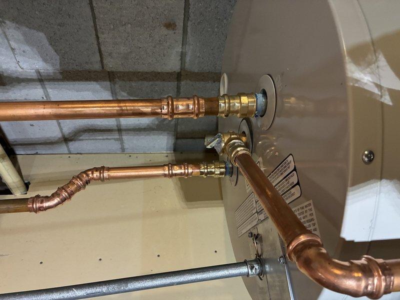 # Plumbing Install - Tanked Water Heater

Completed installation of residential tanked water heater in basement utility area. Installed new cylindrical water heater tank on concrete floor with proper positioning and clearances. Connected hot and cold water supply lines using copper piping with professional compression fittings and joints, forming proper loop configuration at top of unit. Installed brass shut-off valves on both supply lines for isolation and maintenance access. Connected gray galvanized pipe at bottom drain port. Mounted water softener control unit with digital display panel on top of water heater, securing all connections with appropriate fittings. Routed flexible supply hose to top-mounted control device. Verified all copper pipe joints are secure with no visible leaks. Ensured manufacturer labels and safety specifications remain visible and intact. Installation meets code requirements with proper venting configuration and clearances from walls and ceiling. All plumbi