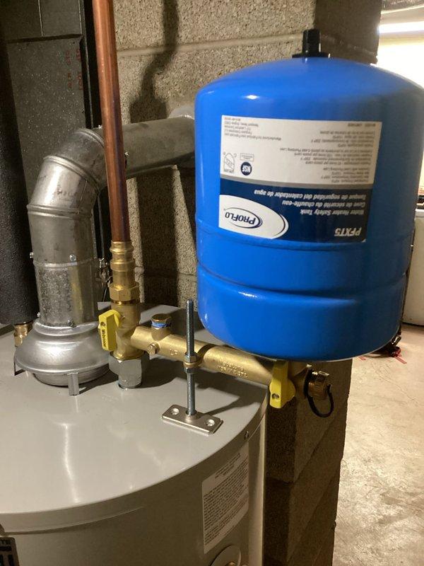Completed installation of residential water heater system with integrated pressure expansion tank in basement mechanical room. Installed white cylindrical water heater unit with proper venting, connected blue expansion tank to copper piping system with brass fittings and shut-off valves, and secured all components to concrete block wall structure with appropriate mounting hardware to ensure code-compliant operation and system pressure regulation.