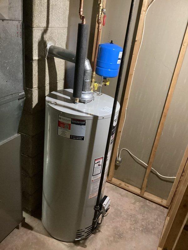 Completed installation of residential water heater system with integrated pressure expansion tank in basement mechanical room. Installed white cylindrical water heater unit with proper venting, connected blue expansion tank to copper piping system with brass fittings and shut-off valves, and secured all components to concrete block wall structure with appropriate mounting hardware to ensure code-compliant operation and system pressure regulation.