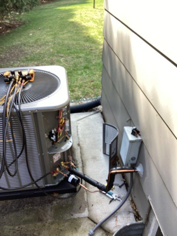 Completed installation of new residential central air conditioning system including outdoor condensing unit and evaporator coil. Mounted gray condensing unit on concrete pad with proper clearances from building structure, installed gray electrical disconnect box on exterior wall per code requirements, and ran refrigerant line set with black foam insulation from outdoor unit through wall penetration to indoor coil location. Connected copper refrigerant lines, electrical wiring, and condensate drain line to complete system integration. Performed vacuum and pressure testing of refrigerant circuit using diagnostic equipment to verify system integrity prior to charging. Successfully commissioned system with all components operating within manufacturer specifications.