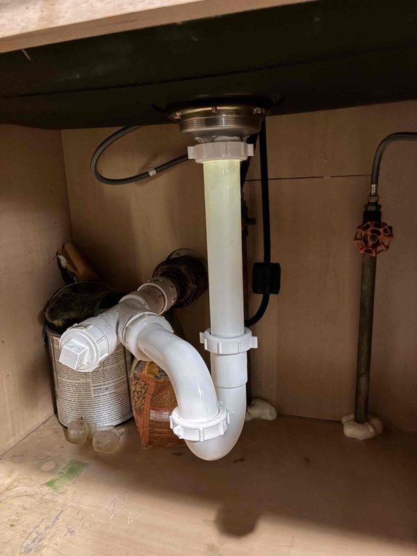 Responded to under-sink plumbing issue involving corroded and deteriorating PVC drain pipe assembly beneath kitchen sink. Upon inspection, identified significant discoloration and corrosion on the vertical drain pipe connecting to the garbage disposal unit, with brownish staining indicating water damage and material breakdown. Replaced the compromised white PVC drain assembly with new components, ensuring proper connection to the existing garbage disposal mounting flange and P-trap configuration. Verified all threaded connections and coupling nuts were properly sealed to prevent future leaks. Tested the complete drain system for proper water flow and confirmed no leaks at connection points.