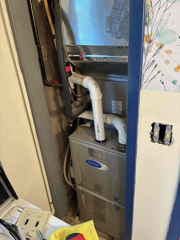 Completed HVAC system installation in residential utility closet featuring Carrier furnace with matching air handler unit. Installation included proper venting configuration with white PVC piping for combustion air and condensate drainage, copper refrigerant line sets with insulation, and electrical connections. All components were mounted and secured within confined mechanical space according to manufacturer specifications and local code requirements.