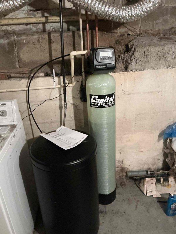 Completed installation of a comprehensive water treatment system in basement utility area, including a black water softener tank and Capital brand water filtration system with digital control head. Installed and connected white PVC piping configuration with shut-off valves, integrated copper supply lines to existing plumbing infrastructure, and established proper drainage connections using flexible corrugated hose. Mounted control components to concrete block wall, completed all electrical connections between units, verified proper system operation, and provided documentation for homeowner reference.