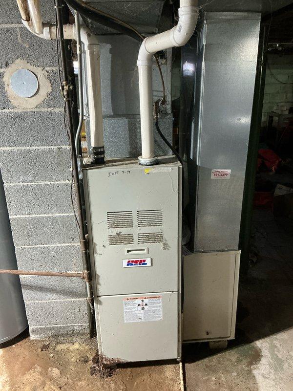 Completed HVAC system assessment in unfinished basement for equipment replacement estimate. Evaluated existing furnace installation, including current white cabinet unit with front ventilation grilles showing age-related wear and discoloration at base. Documented exposed utility infrastructure including white PVC venting system, grey metal ductwork, copper piping with oxidation, black flexible ductwork, and galvanized metal components throughout ceiling joists. Assessed concrete block wall conditions, floor substrate, and clearance requirements for replacement equipment installation in utility space.