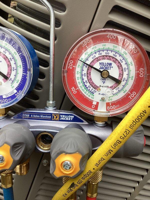 Completed system start-up for newly installed HVAC equipment. Connected manifold gauge set to condenser unit and verified refrigerant pressures across the system, with readings showing proper charge levels on both low-side (blue gauge) and high-side (red gauge) indicators. Measured ambient and system temperatures using digital thermometer, recording 77.1°F to establish baseline operating conditions. All gauges indicated normal operating parameters, confirming successful installation and proper system function upon initial start-up.