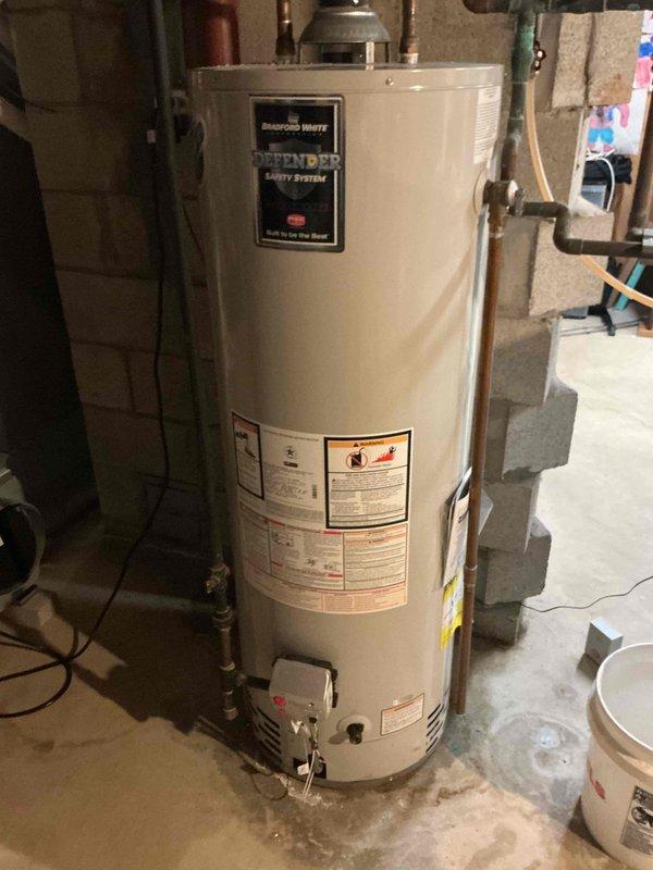 Completed installation inspection and documentation of Bradford White Defender Safety System tanked water heater in residential basement utility area. Unit properly positioned on concrete foundation with appropriate clearances, connected to copper supply lines and metallic flue venting system extending through ceiling joists. Verified all manufacturer safety labels and informational placards present and legible on exterior tank surface. Documented existing copper piping infrastructure showing age-appropriate patina and corrosion, with connections properly secured at ceiling penetration points with insulation wrapping. Confirmed adequate drainage provisions with collection bucket positioned at base of unit, and noted typical concrete floor staining consistent with normal condensation patterns around water heater installation.