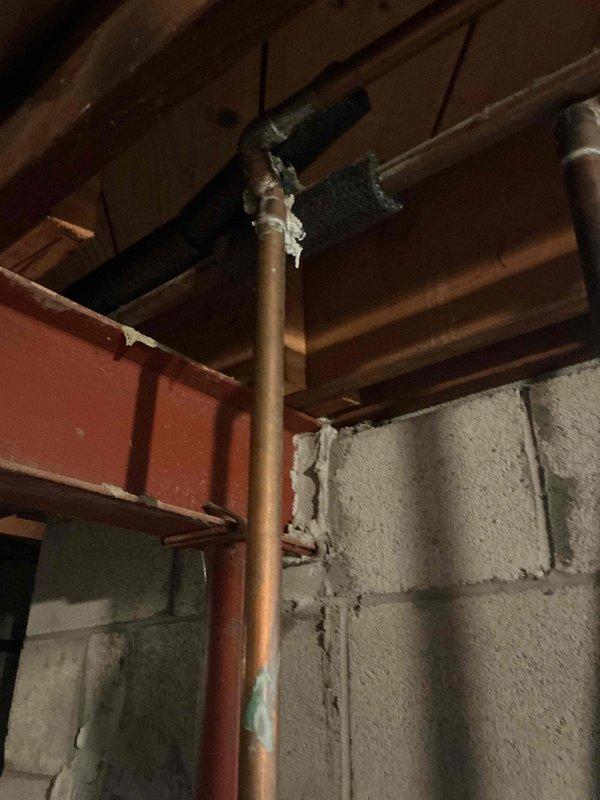 Completed installation inspection and documentation of Bradford White Defender Safety System tanked water heater in residential basement utility area. Unit properly positioned on concrete foundation with appropriate clearances, connected to copper supply lines and metallic flue venting system extending through ceiling joists. Verified all manufacturer safety labels and informational placards present and legible on exterior tank surface. Documented existing copper piping infrastructure showing age-appropriate patina and corrosion, with connections properly secured at ceiling penetration points with insulation wrapping. Confirmed adequate drainage provisions with collection bucket positioned at base of unit, and noted typical concrete floor staining consistent with normal condensation patterns around water heater installation.