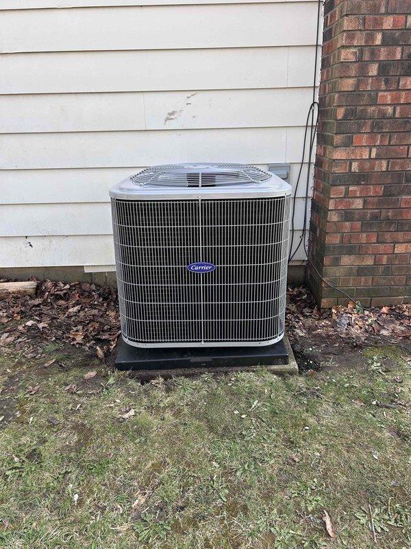 Completed installation of new Carrier air conditioning system with matching evaporator coil at residential property. Installed outdoor condensing unit on elevated platform against exterior wall, ran refrigerant lines and electrical connections through conduit to disconnect box mounted on building siding, and positioned unit for optimal airflow and serviceability. System was pressure tested, evacuated, charged to manufacturer specifications, and verified operational with all safety controls functioning properly.