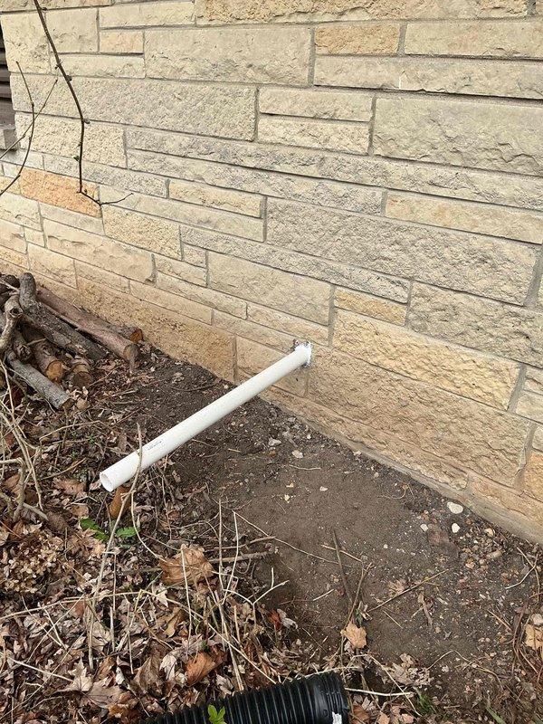 Completed installation of a comprehensive sump pump system in basement, including primary pump unit with circular cover mounted flush in concrete floor, vertical PVC discharge piping secured to foundation wall with mounting brackets, and backup battery power system to ensure continuous operation during power outages. Routed exterior discharge line away from building foundation using white PVC piping to direct water safely from structure. System tested and verified operational with all components properly connected and secured.