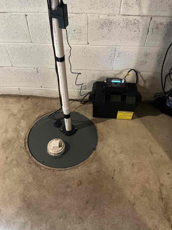 Completed installation of a comprehensive sump pump system in basement, including primary pump unit with circular cover mounted flush in concrete floor, vertical PVC discharge piping secured to foundation wall with mounting brackets, and backup battery power system to ensure continuous operation during power outages. Routed exterior discharge line away from building foundation using white PVC piping to direct water safely from structure. System tested and verified operational with all components properly connected and secured.