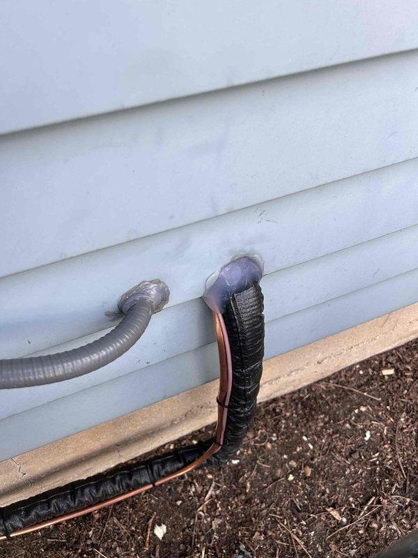 Completed installation of new Lennox ML13XC1 air conditioning system with matching evaporator coil. Installed outdoor condenser unit on ground-level pad adjacent to building exterior, routed refrigerant line set and electrical conduit through exterior wall penetration with proper sealing. Connected copper refrigerant lines with black foam insulation, secured all penetrations through white vinyl siding, and verified proper electrical connections. System tested and operational.