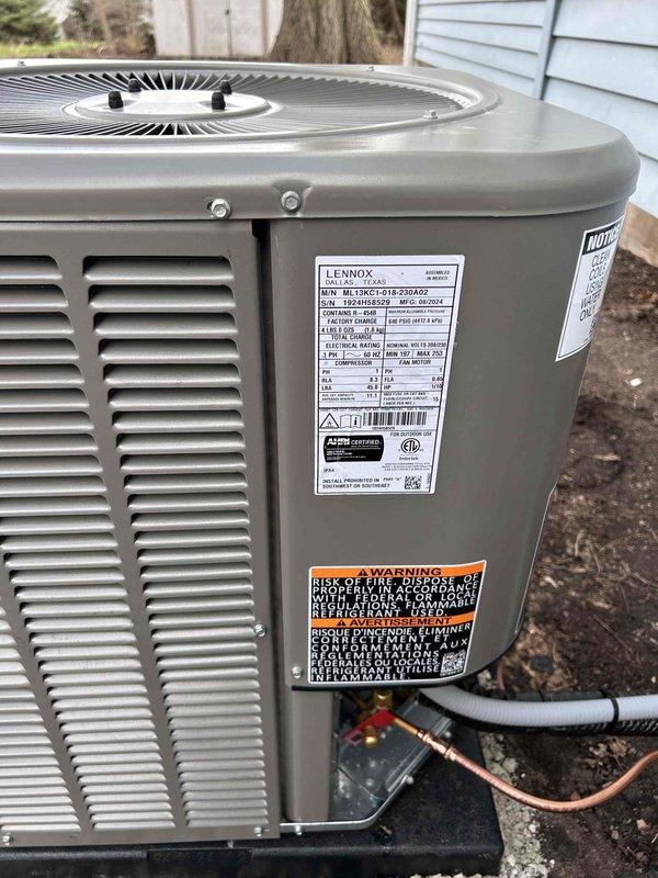 Completed installation of new Lennox ML13XC1 air conditioning system with matching evaporator coil. Installed outdoor condenser unit on ground-level pad adjacent to building exterior, routed refrigerant line set and electrical conduit through exterior wall penetration with proper sealing. Connected copper refrigerant lines with black foam insulation, secured all penetrations through white vinyl siding, and verified proper electrical connections. System tested and operational.