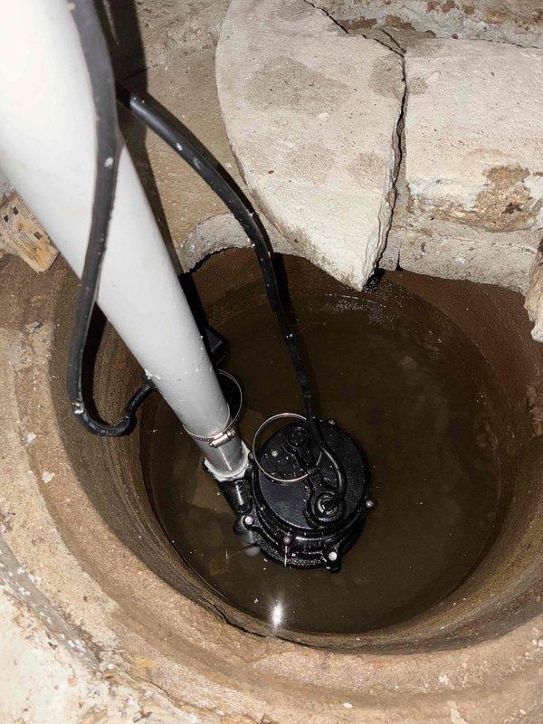Completed installation of residential sump pump system in basement floor basin. Installed black submersible pump unit in existing concrete pit with white PVC discharge piping and electrical connection, secured with metal hose clamp. System tested and operational for basement water management and flood prevention.