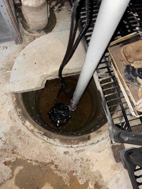 Completed installation of residential sump pump system in basement floor basin. Installed black submersible pump unit in existing concrete pit with white PVC discharge piping and electrical connection, secured with metal hose clamp. System tested and operational for basement water management and flood prevention.