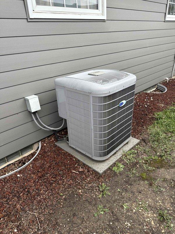 Conducted comprehensive assessment of existing HVAC system for replacement estimate, including outdoor air conditioning condenser unit and indoor furnace system. Evaluated current 2-ton AC condenser positioned on concrete pad against exterior wall with gray electrical conduit connections and documented condition of unit housing, heat exchange coils, and protective grille assembly. Inspected basement furnace installation including ductwork configuration, metal cabinet construction, refrigerant line routing, and electrical connections. Photographed both indoor and outdoor equipment to document existing system specifications, installation methods, and spatial constraints for replacement planning. Assessed concrete pad condition, electrical service routing along vinyl siding, and clearance requirements around outdoor unit within mulched landscaping area. Reviewed basement mechanical room layout including exposed floor joists, available workspace, and existing duct transitions to determine 