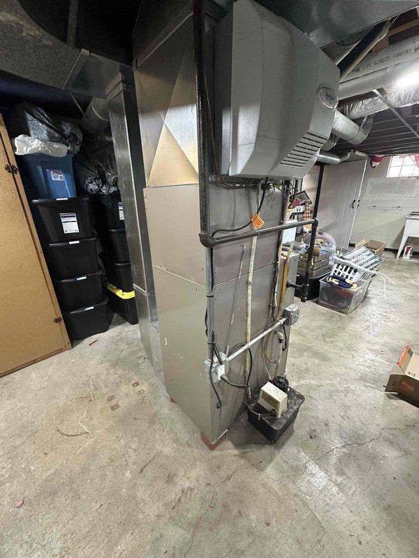 Conducted comprehensive assessment of existing HVAC system for replacement estimate, including outdoor air conditioning condenser unit and indoor furnace system. Evaluated current 2-ton AC condenser positioned on concrete pad against exterior wall with gray electrical conduit connections and documented condition of unit housing, heat exchange coils, and protective grille assembly. Inspected basement furnace installation including ductwork configuration, metal cabinet construction, refrigerant line routing, and electrical connections. Photographed both indoor and outdoor equipment to document existing system specifications, installation methods, and spatial constraints for replacement planning. Assessed concrete pad condition, electrical service routing along vinyl siding, and clearance requirements around outdoor unit within mulched landscaping area. Reviewed basement mechanical room layout including exposed floor joists, available workspace, and existing duct transitions to determine 