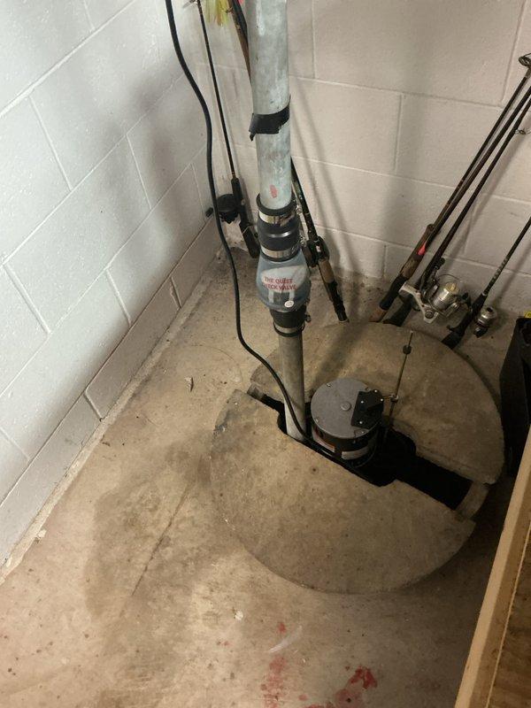 Performed inspection and assessment of existing sump pump system in basement utility area. Documented current condition of pump assembly, discharge piping, and basin installation. Observed evidence of previous water accumulation and moisture issues based on floor staining patterns. Verified sump pump operation, electrical connections, and discharge pipe mounting. Checked basin positioning within floor pit and confirmed proper drainage configuration. Photographed system components including pump mechanism, vertical discharge assembly with wall-mounted brackets, and associated plumbing connections for service records.
