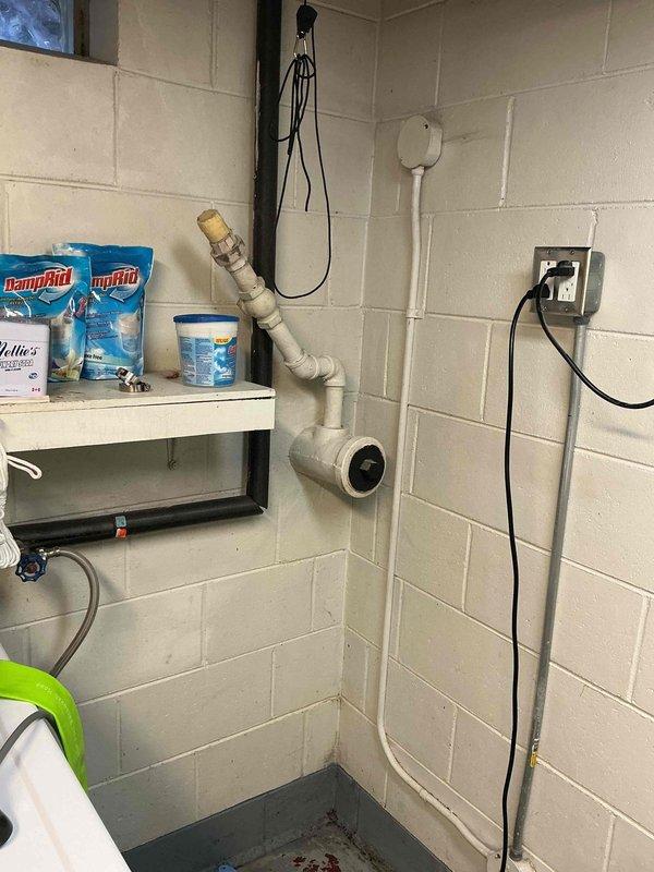 Performed sump pump inspection and assessment in basement utility area. Evaluated existing blue submersible sump pump installed in below-grade concrete pit with brick-lined walls. Verified pump positioning and operational setup within basin. Documented electrical connections and power supply routing, including pump cord configuration. Assessed basement moisture conditions, noting rust staining on concrete floor indicating previous water intrusion or ongoing moisture issues. Confirmed proper drainage pipe assembly consisting of white PVC discharge piping with multiple elbow joints forming vertical wall-mounted configuration for water evacuation. System appears functional with appropriate components in place for groundwater management and basement flood prevention.