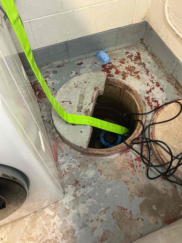 Performed sump pump inspection and assessment in basement utility area. Evaluated existing blue submersible sump pump installed in below-grade concrete pit with brick-lined walls. Verified pump positioning and operational setup within basin. Documented electrical connections and power supply routing, including pump cord configuration. Assessed basement moisture conditions, noting rust staining on concrete floor indicating previous water intrusion or ongoing moisture issues. Confirmed proper drainage pipe assembly consisting of white PVC discharge piping with multiple elbow joints forming vertical wall-mounted configuration for water evacuation. System appears functional with appropriate components in place for groundwater management and basement flood prevention.