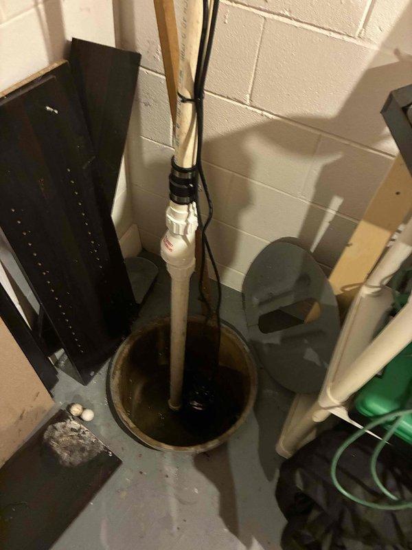 Completed plumbing estimate for existing sump pump system in basement utility area. Evaluated current sump pump installation including basin condition, discharge piping configuration, pump operation, and electrical connections. Documented system setup with vertical PVC discharge pipe mounted to wall with proper securing, pump placement within circular basin, and water level conditions at time of inspection to provide comprehensive assessment for potential service or replacement recommendations.