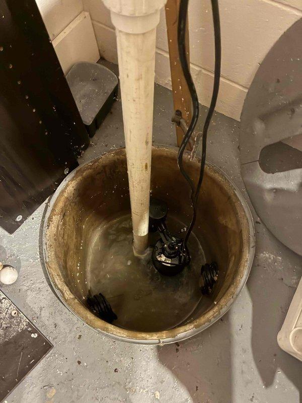 Completed plumbing estimate for existing sump pump system in basement utility area. Evaluated current sump pump installation including basin condition, discharge piping configuration, pump operation, and electrical connections. Documented system setup with vertical PVC discharge pipe mounted to wall with proper securing, pump placement within circular basin, and water level conditions at time of inspection to provide comprehensive assessment for potential service or replacement recommendations.