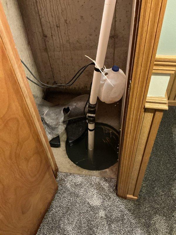 Performed comprehensive inspection of existing sump pump system located in basement. Evaluated sump basin condition, noting water accumulation with visible sediment and mineral content present in basin. Inspected vertical PVC discharge pipe installation, verifying proper mounting with securing brackets and clamps. Assessed electrical connections and cord routing from pump unit. Examined basin perimeter and surrounding concrete floor, documenting debris accumulation and general wear patterns. Verified pump accessibility and basin cover components. System remains operational for continued water management and flood prevention purposes.