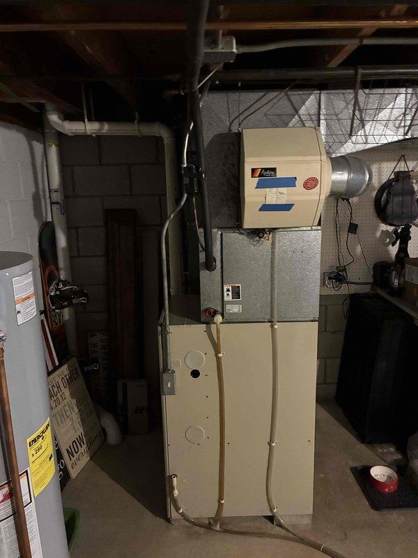 Completed estimate for replacement of existing gas furnace and air conditioning system in basement mechanical room. Both units currently operational but exhibiting age-related noise issues. Assessed existing beige cabinet-style furnace with associated ductwork, refrigerant lines, and electrical connections mounted on concrete block wall. Evaluated air handler configuration, copper tubing connections, and existing control panel installation. Photographed current equipment layout including positioning relative to water heater and exposed ceiling joists to determine replacement specifications and installation requirements.