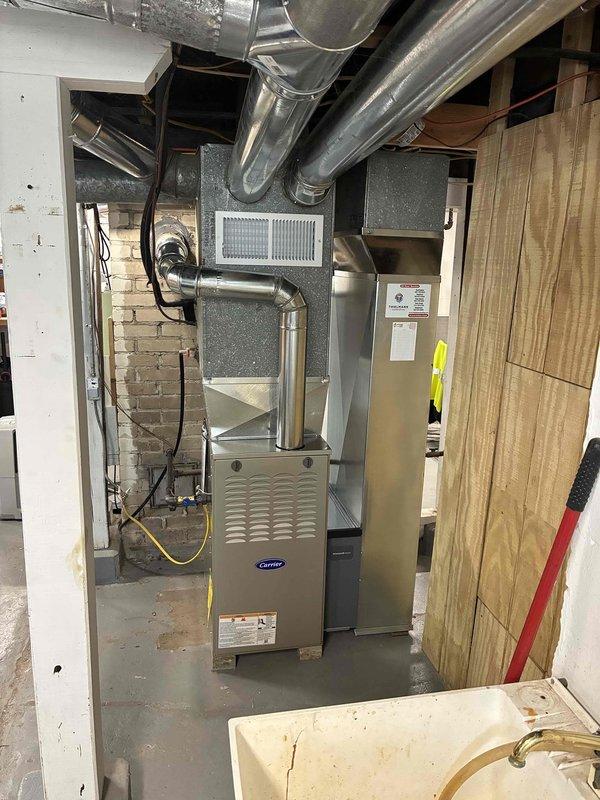 Completed installation of new Carrier furnace unit in basement mechanical room with comprehensive ductwork integration. Installed metallic ductwork system throughout ceiling area, including main trunk lines and branch connections to distribute conditioned air throughout the structure. Configured electrical connections with proper conduit routing and junction box mounting to exposed masonry wall, ensuring code-compliant power supply to heating unit. Positioned furnace on concrete floor pad and connected to existing duct network, with all ventilation grills and safety labels properly installed on unit exterior.