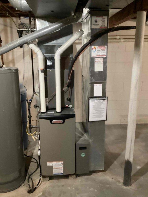 Completed residential furnace installation in basement utility area. New gray furnace unit installed with proper PVC venting routed through ceiling joists for exhaust and combustion air intake. Unit positioned adjacent to existing water heater with appropriate clearances maintained for service access and code compliance. All manufacturer labels and safety documentation affixed to equipment. Electrical connections completed and ductwork integrated with existing HVAC distribution system. Installation includes standard operational and safety features per manufacturer specifications. System tested and verified operational upon completion.