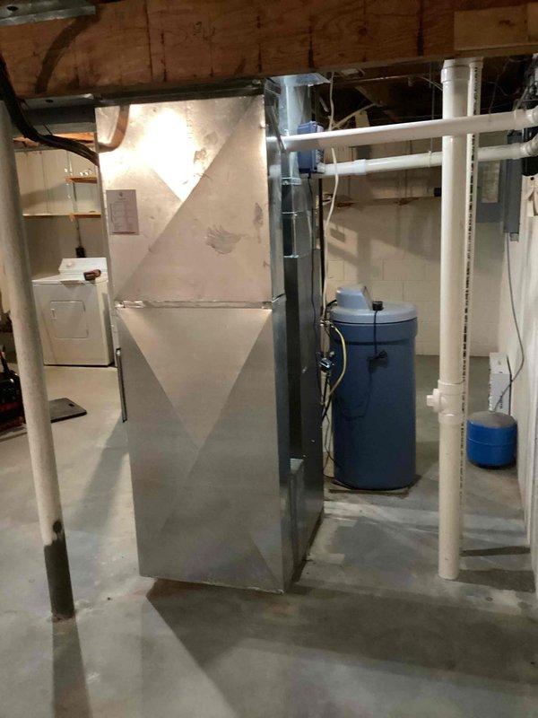 Completed residential furnace installation in basement utility area. New gray furnace unit installed with proper PVC venting routed through ceiling joists for exhaust and combustion air intake. Unit positioned adjacent to existing water heater with appropriate clearances maintained for service access and code compliance. All manufacturer labels and safety documentation affixed to equipment. Electrical connections completed and ductwork integrated with existing HVAC distribution system. Installation includes standard operational and safety features per manufacturer specifications. System tested and verified operational upon completion.