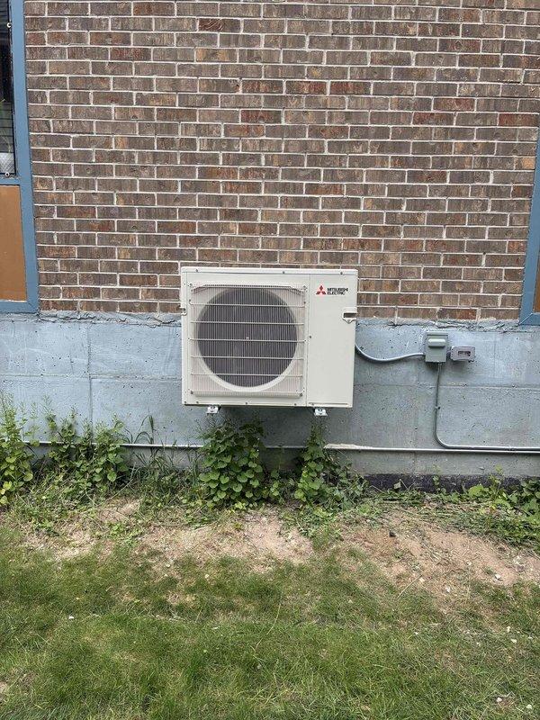 Completed installation of Mitsubishi Electric mini-split system including exterior condenser unit mounted on concrete foundation with proper electrical disconnect and interior wall-mounted air handler in living room. All refrigerant lines were run through exterior wall, system was tested and verified operational, and installation meets manufacturer specifications for residential heating and cooling application.