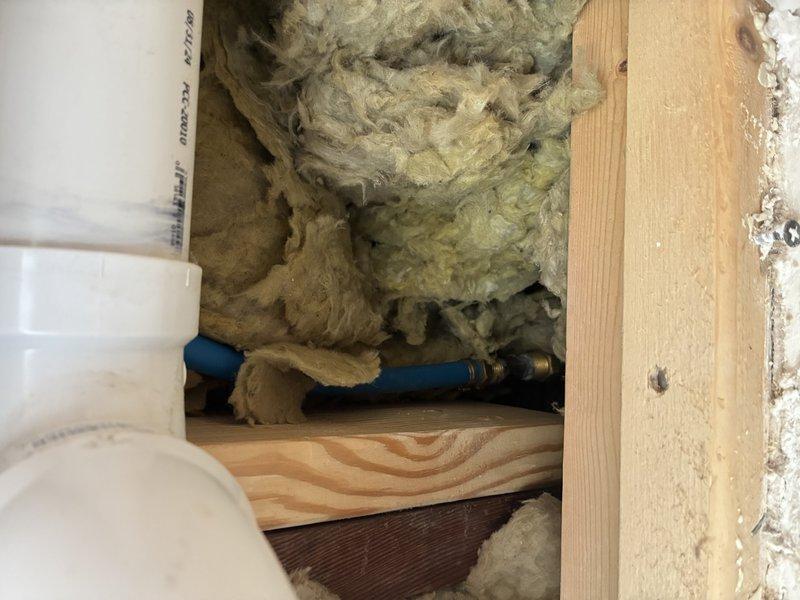Responded to service call regarding malfunctioning outdoor hose bib with suspected pipe damage. Upon investigation, accessed interior wall cavity to expose supply line running to exterior spigot. Identified failed or damaged piping requiring replacement. Created access opening through interior wall to reach affected plumbing section. Observed existing white PVC supply line with brass fittings and blue water line running through floor/wall cavity between framing members. Assessed condition of insulation and structural framing during repair work. Documented conditions via photographs for service records.